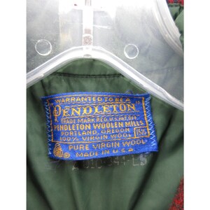 Puede incluir: El interior de una prenda de color verde oscuro con una etiqueta de tela azul rectangular que presenta el texto bordado en amarillo: Pendleton, Warranted to be a Pendleton, Pendleton Woolen Mills, Portland, Oregon, 100% Virgin Wool, Pure Virgin Wool, Made in U.S.A.