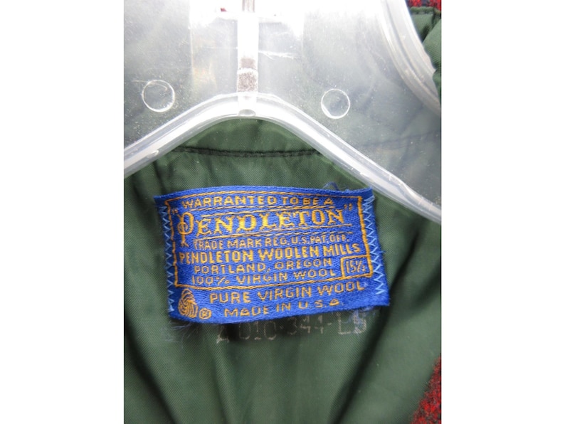 Puede incluir: El interior de una prenda de color verde oscuro con una etiqueta de tela azul rectangular que presenta el texto bordado en amarillo: Pendleton, Warranted to be a Pendleton, Pendleton Woolen Mills, Portland, Oregon, 100% Virgin Wool, Pure Virgin Wool, Made in U.S.A.
