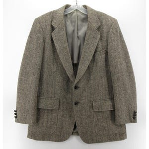 May include: A brown tweed blazer with a herringbone pattern. The jacket features two front pockets, a chest pocket, and black buttons. It is displayed on a hanger against a white backdrop.
