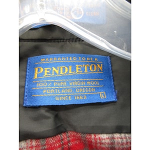 May include: A blue rectangular clothing label with yellow text reading Warranted To Be A Pendleton, 100% Pure Virgin Wool, Portland, Oregon, Since 1863, and the size L. The label is attached to a black garment with a red and grey plaid lining.
