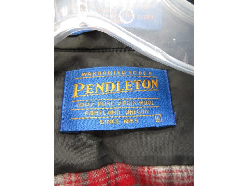 May include: A blue rectangular clothing label with yellow text reading Warranted To Be A Pendleton, 100% Pure Virgin Wool, Portland, Oregon, Since 1863, and the size L. The label is attached to a black garment with a red and grey plaid lining.