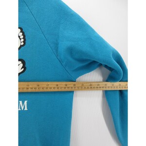 May include: A bright blue long-sleeve sweatshirt featuring a white graphic of feet and the letter M. A wooden ruler is placed across the chest, showing a measurement of approximately 14 inches from the armpit seam to the center of the garment.