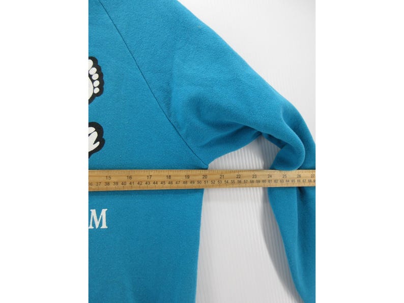 May include: A bright blue long-sleeve sweatshirt featuring a white graphic of feet and the letter M. A wooden ruler is placed across the chest, showing a measurement of approximately 14 inches from the armpit seam to the center of the garment.