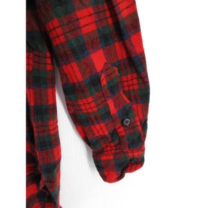 Puede incluir: Una camisa de franela de manga larga con un patr&oacute;n de cuadros en rojo, verde oscuro y negro. La manga incluye un pu&ntilde;o abotonado con un bot&oacute;n negro visible, y la tela presenta una textura suave y cepillada en toda la prenda.