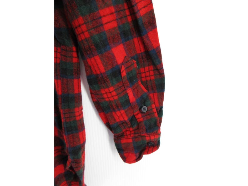 Puede incluir: Una camisa de franela de manga larga con un patr&oacute;n de cuadros en rojo, verde oscuro y negro. La manga incluye un pu&ntilde;o abotonado con un bot&oacute;n negro visible, y la tela presenta una textura suave y cepillada en toda la prenda.