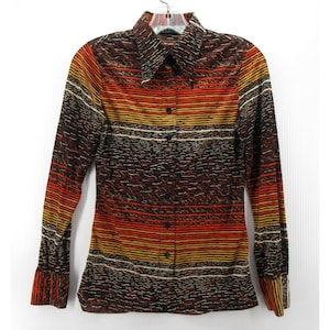 Peut inclure: Une chemise à manches longues avec un design rétro. La chemise présente des rayures horizontales dans les tons marron, orange, jaune et blanc. Elle a un col pointu et des poignets boutonnés. La chemise est suspendue à un cintre en plastique transparent.