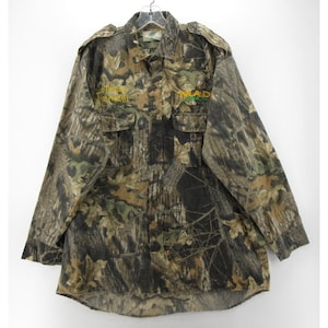 Puede incluir: Camisa de camuflaje con botones y mangas largas. La camisa presenta un patr&oacute;n de camuflaje de bosque en tonos marrones, verdes y beige. Tiene dos bolsillos en el pecho y el texto "MAD" en amarillo y verde en la parte delantera.