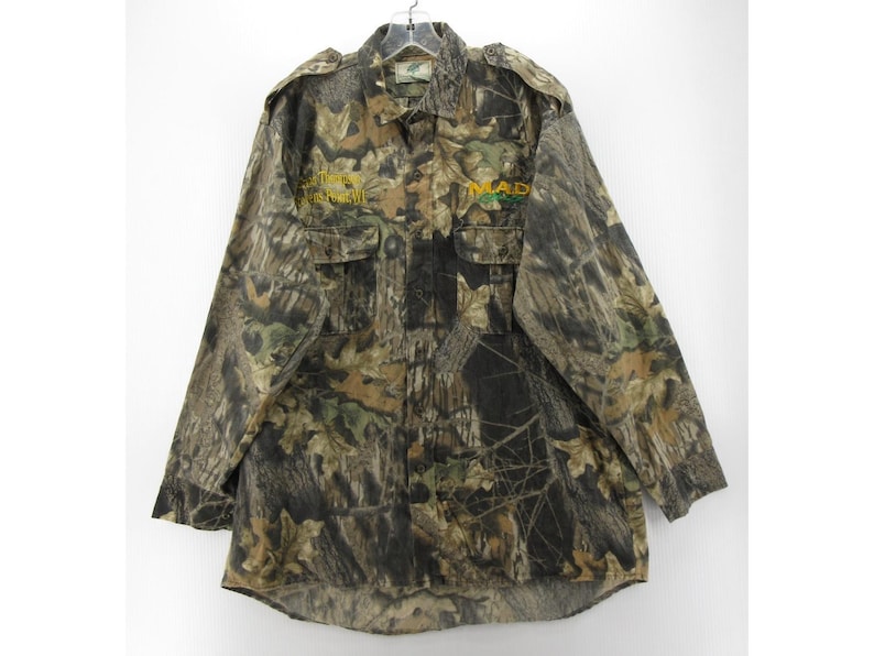 Puede incluir: Camisa de camuflaje con botones y mangas largas. La camisa presenta un patr&oacute;n de camuflaje de bosque en tonos marrones, verdes y beige. Tiene dos bolsillos en el pecho y el texto "MAD" en amarillo y verde en la parte delantera.