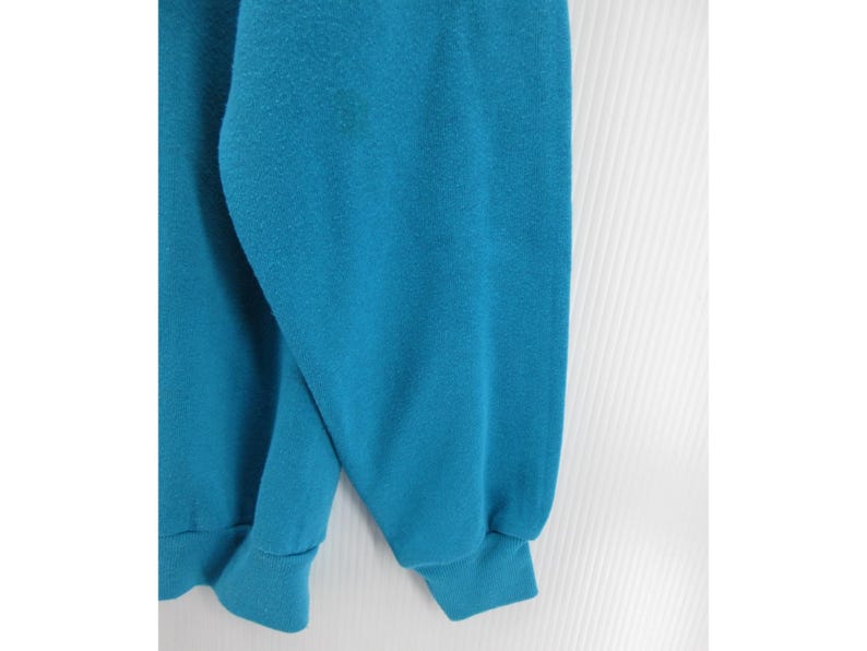 May include: A solid teal long-sleeve sweatshirt featuring a crew neckline and ribbed cuffs at the wrists and hem. The garment is made of a soft, fleece-like fabric with a consistent, vibrant blue-green color throughout the entire visible surface.