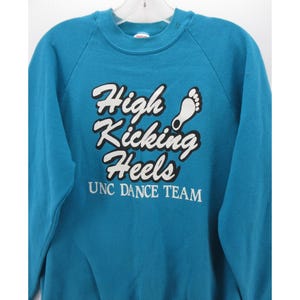 May include: A teal crewneck sweatshirt featuring the white text "High Kicking Heels" with a black outline and a white footprint graphic. Below the main text, the words "UNC DANCE TEAM" are printed in white. The garment has long sleeves.