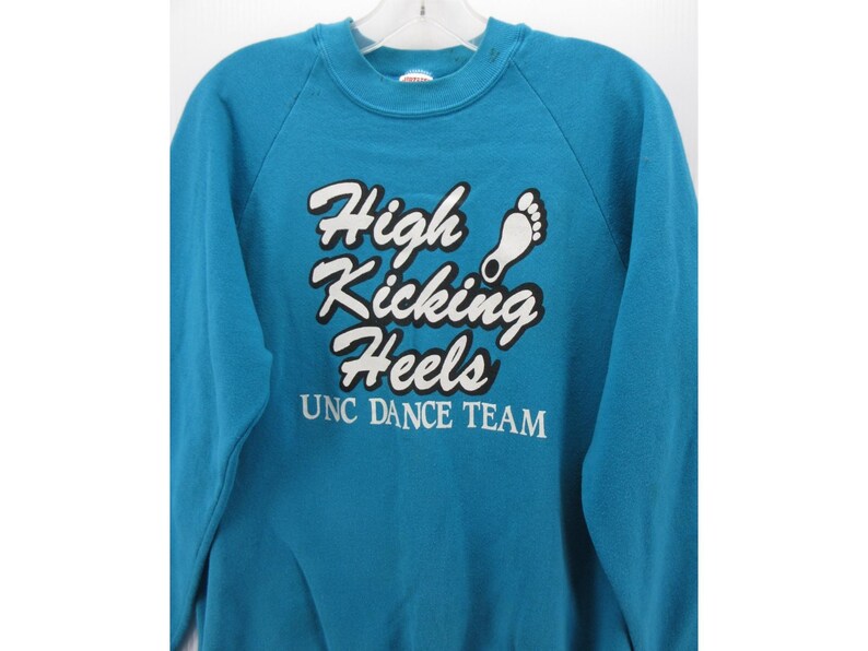 May include: A teal crewneck sweatshirt featuring the white text "High Kicking Heels" with a black outline and a white footprint graphic. Below the main text, the words "UNC DANCE TEAM" are printed in white. The garment has long sleeves.