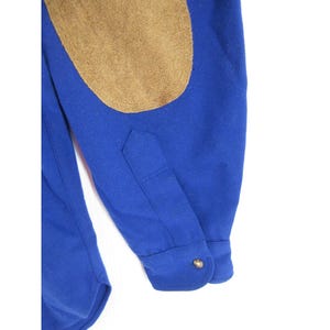 May include: A long-sleeved royal blue shirt featuring a tan suede elbow patch on the sleeve. The sleeve includes a buttoned cuff with a single metallic button and a pointed placket detail. The fabric appears to be a solid, smooth material.