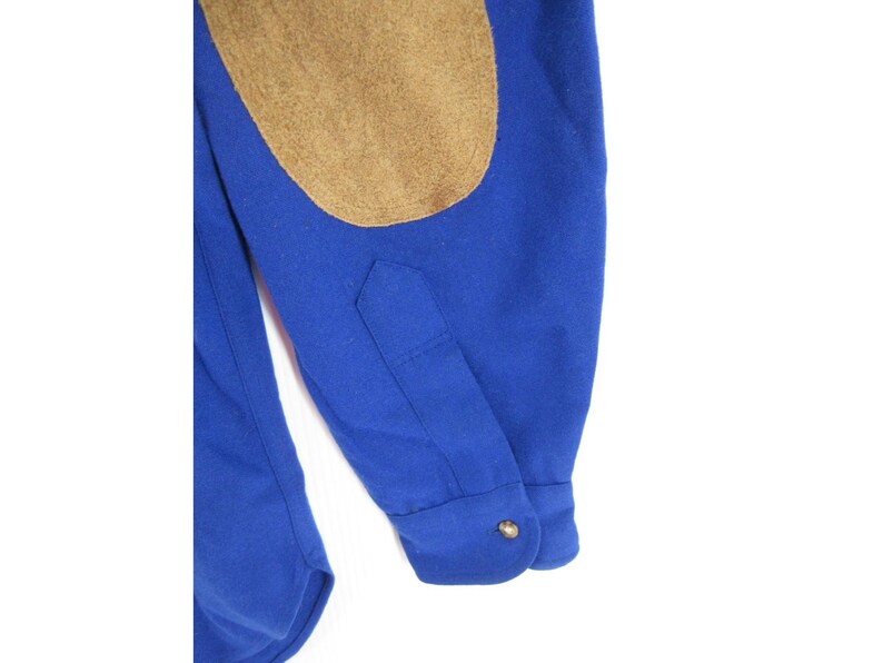 May include: A long-sleeved royal blue shirt featuring a tan suede elbow patch on the sleeve. The sleeve includes a buttoned cuff with a single metallic button and a pointed placket detail. The fabric appears to be a solid, smooth material.