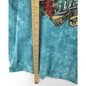 May include: A turquoise tie-dye t-shirt with a graphic featuring red roses and the text "DE" above a banner that reads "SINCE". A wooden ruler is placed vertically along the side of the shirt.