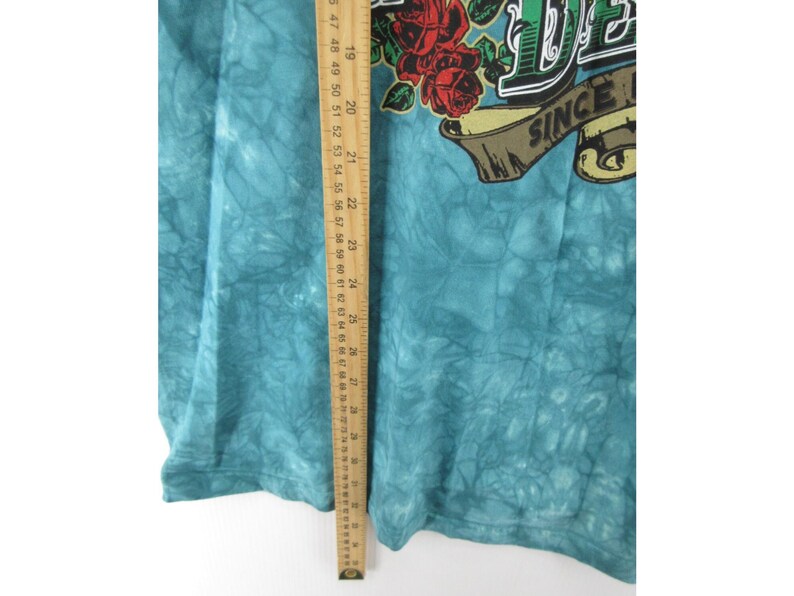 May include: A turquoise tie-dye t-shirt with a graphic featuring red roses and the text "DE" above a banner that reads "SINCE". A wooden ruler is placed vertically along the side of the shirt.