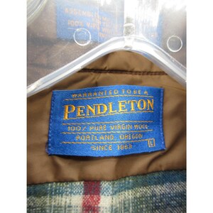 May include: A brown garment featuring a blue rectangular label with yellow text that reads Warranted To Be A Pendleton, 100% Pure Virgin Wool, Portland, Oregon, Since 1863, with a size L indicator. The garment has a plaid lining with red, blue, and white.