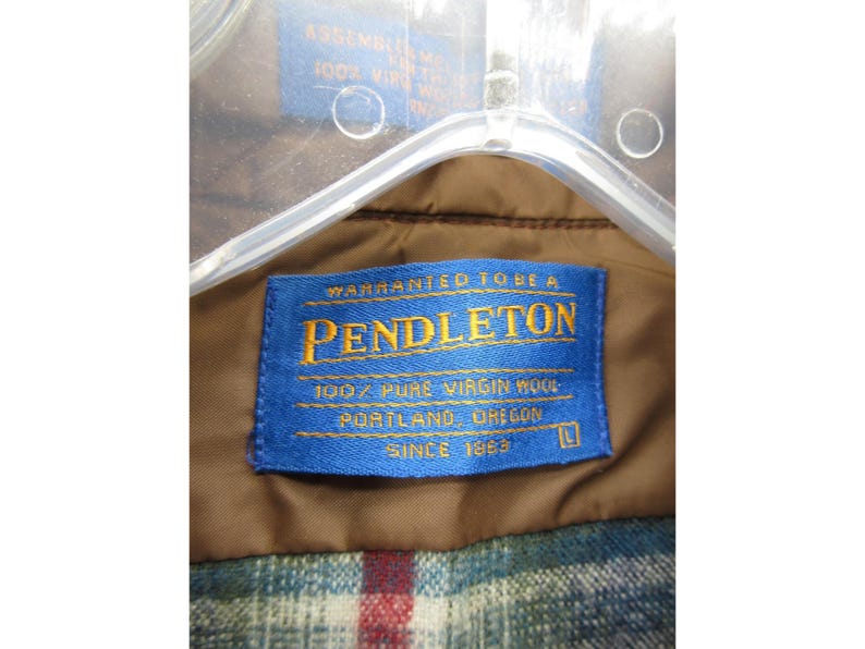 May include: A brown garment featuring a blue rectangular label with yellow text that reads Warranted To Be A Pendleton, 100% Pure Virgin Wool, Portland, Oregon, Since 1863, with a size L indicator. The garment has a plaid lining with red, blue, and white.