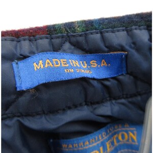 May include: A dark blue quilted interior lining of a garment featuring a blue fabric tag with the text MADE IN U.S.A. and RN 20605 in gold lettering. Above the tag is a section of dark multicolored plaid wool fabric with a textured surface.