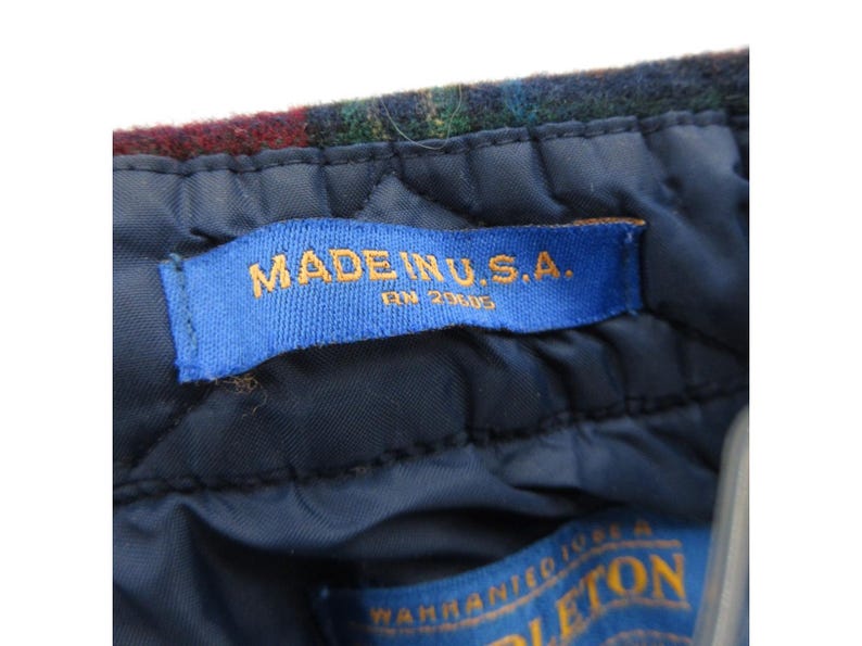 May include: A dark blue quilted interior lining of a garment featuring a blue fabric tag with the text MADE IN U.S.A. and RN 20605 in gold lettering. Above the tag is a section of dark multicolored plaid wool fabric with a textured surface.