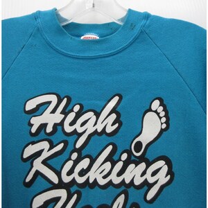 May include: A teal crewneck sweatshirt featuring the white text High Kicking Heels with a black outline and a white graphic of a footprint. The garment has a ribbed collar and long sleeves, with a Jerzees brand label visible at the inner neckline.