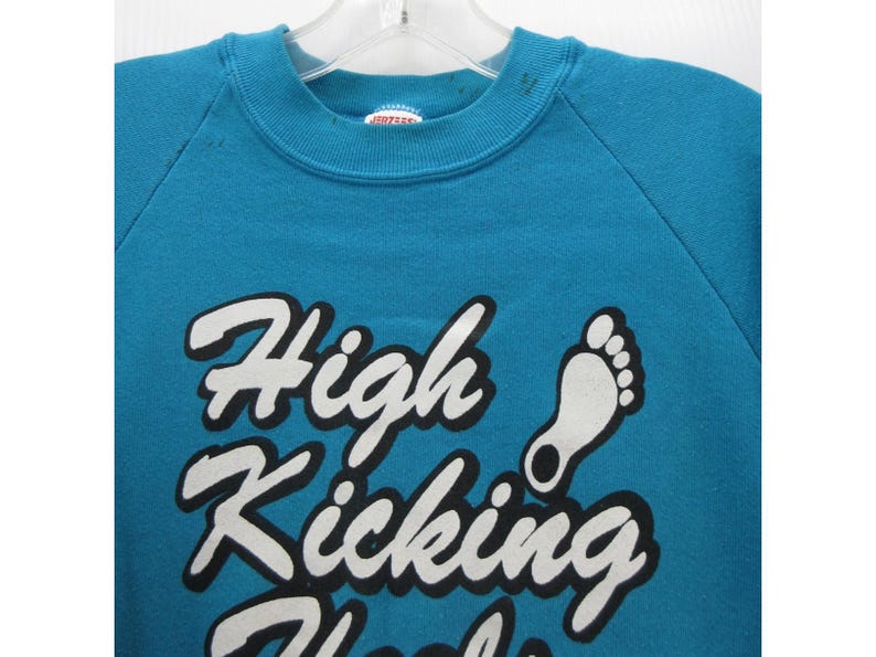 May include: A teal crewneck sweatshirt featuring the white text High Kicking Heels with a black outline and a white graphic of a footprint. The garment has a ribbed collar and long sleeves, with a Jerzees brand label visible at the inner neckline.