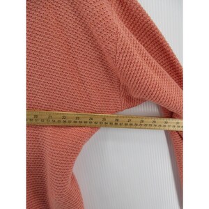 May include: A coral-colored knit sweater with a textured pattern. A wooden ruler is placed across the sweater, measuring approximately 30 inches. The sweater's fabric appears soft and warm, suitable for cooler weather.