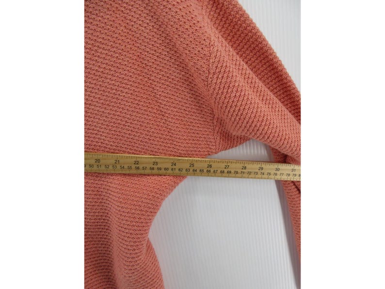 May include: A coral-colored knit sweater with a textured pattern. A wooden ruler is placed across the sweater, measuring approximately 30 inches. The sweater's fabric appears soft and warm, suitable for cooler weather.