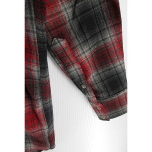 May include: A long-sleeved flannel shirt featuring a red, grey, and black plaid pattern. The sleeve includes a buttoned cuff with three small metallic buttons visible along the placket and cuff edge, showcasing the fabric texture and weave.
