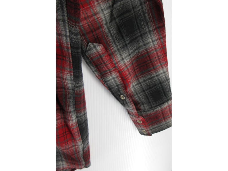 May include: A long-sleeved flannel shirt featuring a red, grey, and black plaid pattern. The sleeve includes a buttoned cuff with three small metallic buttons visible along the placket and cuff edge, showcasing the fabric texture and weave.