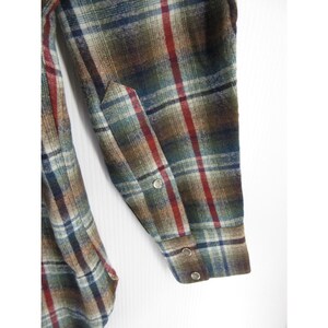 May include: A long-sleeved shirt featuring a plaid pattern in shades of brown, green, blue, and red. The sleeve includes a pointed cuff detail with three metallic snap buttons, showcasing a textured fabric weave throughout the garment.