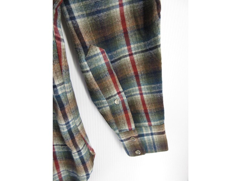 May include: A long-sleeved shirt featuring a plaid pattern in shades of brown, green, blue, and red. The sleeve includes a pointed cuff detail with three metallic snap buttons, showcasing a textured fabric weave throughout the garment.