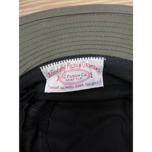 Op de afbeelding: Close-up van de binnenkant van een olijfgroene Filson vissershoed. De hoed heeft een zwarte binnenkant en een wit label met rode tekst: "GENUINE FILSON GARMENT, C. FILSON CO. SEATTLE, MIGHT AS WELL HAVE THE BEST."