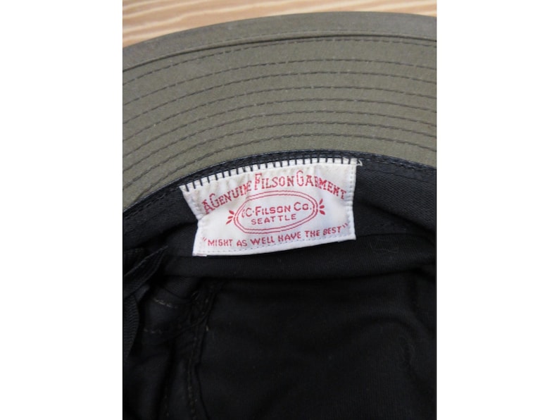 Op de afbeelding: Close-up van de binnenkant van een olijfgroene Filson vissershoed. De hoed heeft een zwarte binnenkant en een wit label met rode tekst: "GENUINE FILSON GARMENT, C. FILSON CO. SEATTLE, MIGHT AS WELL HAVE THE BEST."