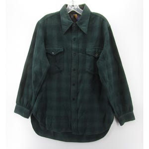 Peut inclure: Une chemise en flanelle vert foncé avec un motif à carreaux. La chemise à manches longues a deux poches de poitrine et une fermeture boutonnée sur le devant. Le col est d'une nuance de vert plus foncée. La chemise est suspendue à un cintre sur fond blanc.