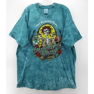 May include: Teal tie-dye t-shirt featuring a graphic of a skeleton with roses and the text "Grateful Dead" and "San Francisco's Own Since 1965". The shirt has short sleeves and a crew neck.