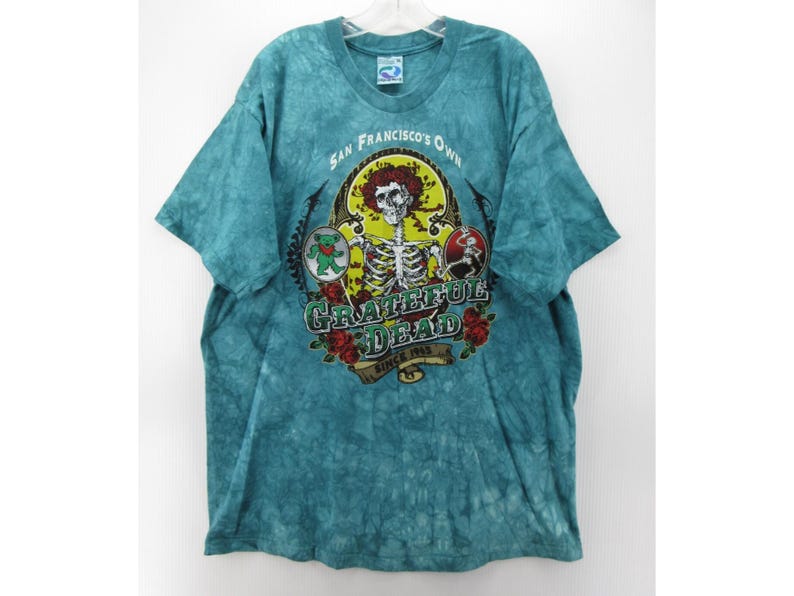 May include: Teal tie-dye t-shirt featuring a graphic of a skeleton with roses and the text "Grateful Dead" and "San Francisco's Own Since 1965". The shirt has short sleeves and a crew neck.