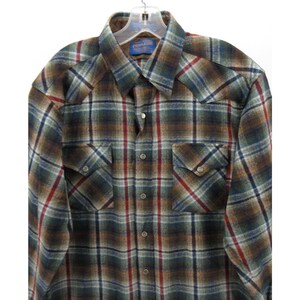 May include: A long-sleeve button-down shirt featuring a plaid pattern in shades of brown, dark green, blue, and red. The shirt includes a pointed collar, two chest pockets with buttoned flaps, and metallic snap buttons down the front placket.