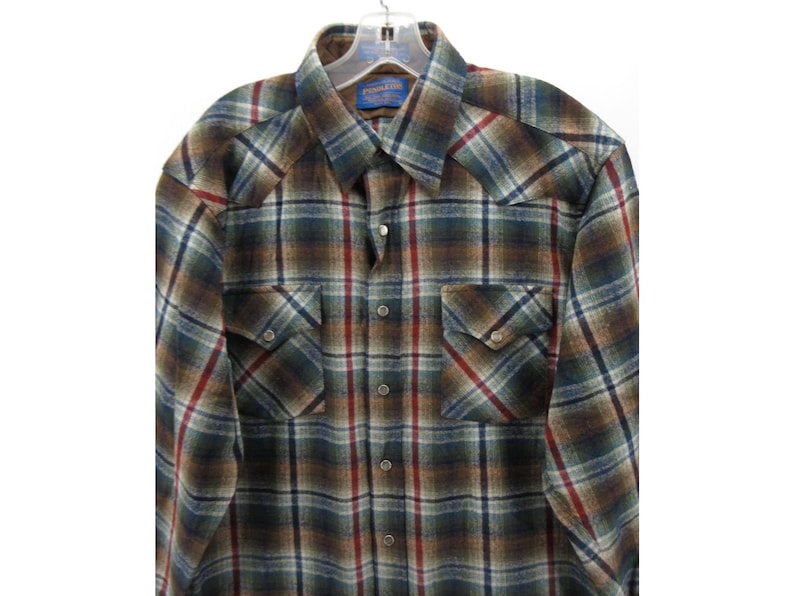 May include: A long-sleeve button-down shirt featuring a plaid pattern in shades of brown, dark green, blue, and red. The shirt includes a pointed collar, two chest pockets with buttoned flaps, and metallic snap buttons down the front placket.