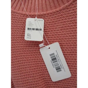May include: Close-up of a coral-colored knit sweater. The sweater has a textured pattern and a round neckline. Attached tags indicate size XL.