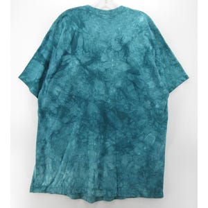 May include: A teal tie-dye t-shirt with short sleeves. The shirt is hanging on a white hanger against a white background. The fabric has a mottled pattern of varying shades of teal.