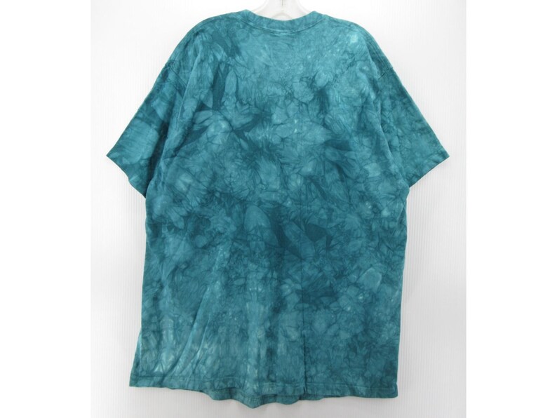 May include: A teal tie-dye t-shirt with short sleeves. The shirt is hanging on a white hanger against a white background. The fabric has a mottled pattern of varying shades of teal.