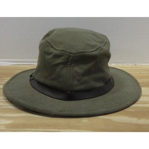 Op de afbeelding: Olijfgroene hoed met een brede rand en een donkerbruine band. De hoed heeft een gestructureerde kroon en een casual stijl. De hoed ligt op een houten oppervlak.