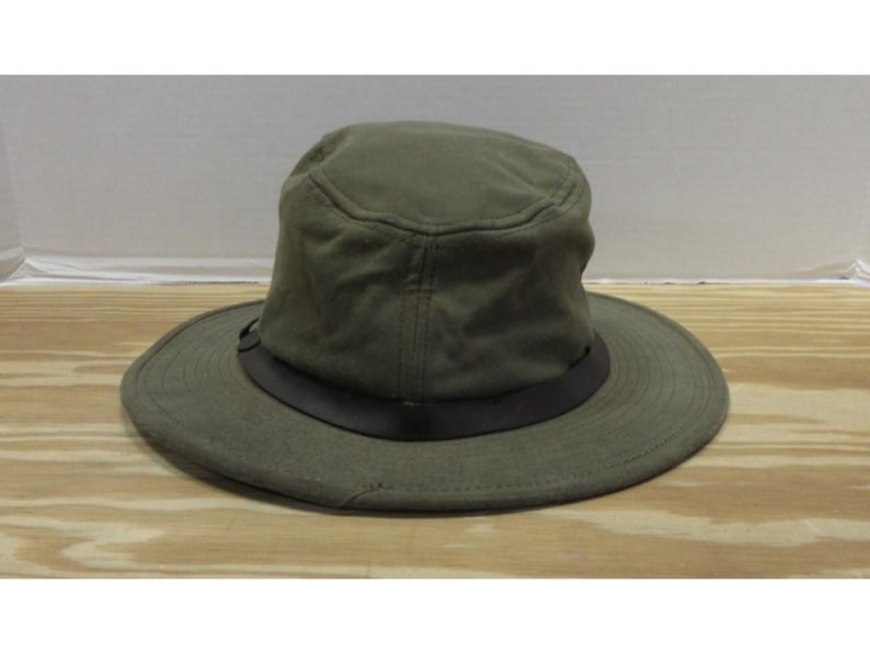 Op de afbeelding: Olijfgroene hoed met een brede rand en een donkerbruine band. De hoed heeft een gestructureerde kroon en een casual stijl. De hoed ligt op een houten oppervlak.