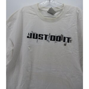 May include: Off-white t-shirt with the phrase "JUST DO IT" printed in black, with gray accents. The shirt has a crew neck and short sleeves. The fabric appears to be slightly wrinkled.