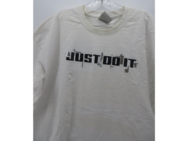May include: Off-white t-shirt with the phrase "JUST DO IT" printed in black, with gray accents. The shirt has a crew neck and short sleeves. The fabric appears to be slightly wrinkled.