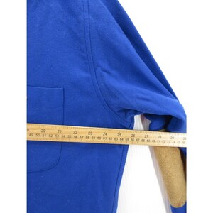 May include: A royal blue long-sleeve shirt featuring a chest pocket and tan elbow patches. A wooden ruler is positioned across the garment, showing a measurement of approximately 28 inches from the center to the sleeve edge.