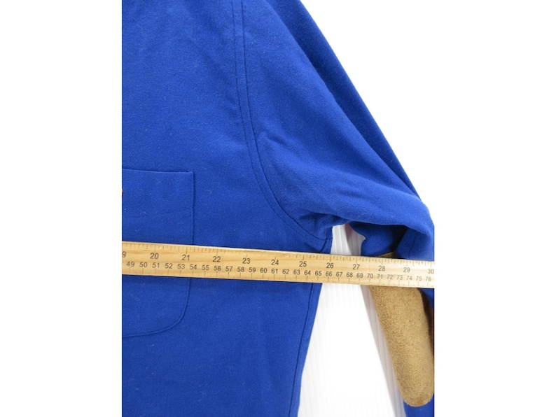 May include: A royal blue long-sleeve shirt featuring a chest pocket and tan elbow patches. A wooden ruler is positioned across the garment, showing a measurement of approximately 28 inches from the center to the sleeve edge.