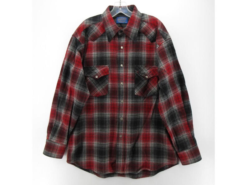 May include: A long-sleeved button-down shirt featuring a red, black and grey plaid pattern. The shirt includes a pointed collar, two chest pockets with buttoned flaps, and metallic snap buttons running down the front placket and on the cuffs.