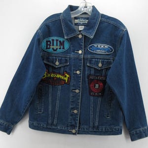 May include: A medium blue denim jacket featuring a classic collar, long sleeves, and a button-up front. The jacket is decorated with four distinct embroidered patches, including text that reads BUM, BUM EQUIPMENT, and AUTHENTIC BUM EQUIPMENT DENIM.