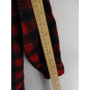 Puede incluir: Una camisa de franela de manga larga con un patr&oacute;n de cuadros rojos y negros. Una regla de madera est&aacute; colocada verticalmente a lo largo de la manga, mostrando medidas de aproximadamente 33 cm a 56 cm para indicar la longitud.